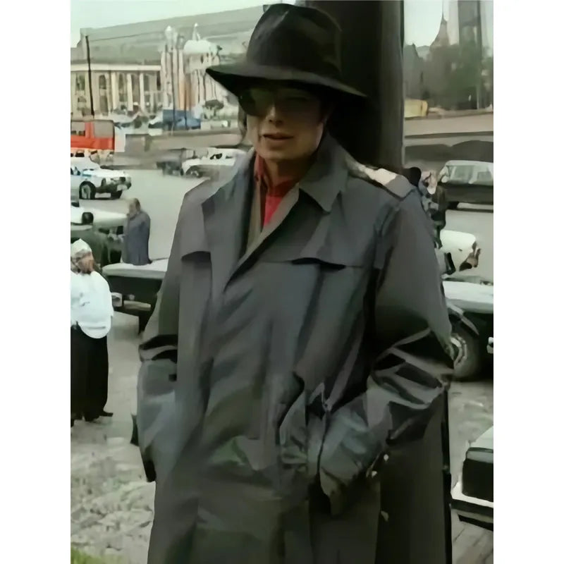 Person wearing a gray coat and hat in an outdoor setting with vehicles and buildings in the background.