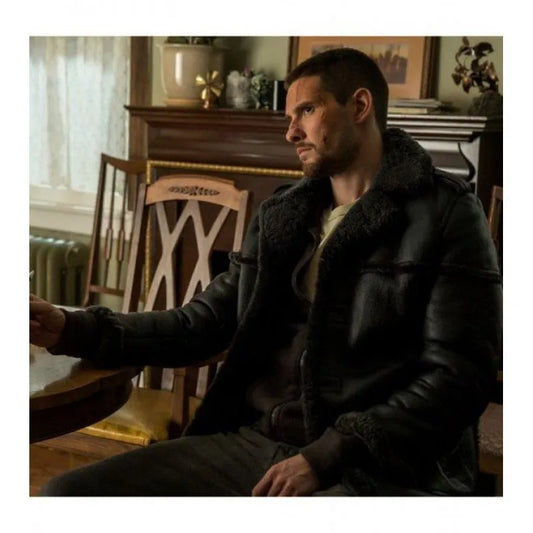 A man wearing a black leather aviator jacket with dark shearling lining sits in a dimly lit, vintage-style room.