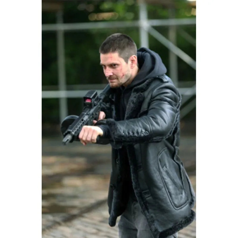A man wearing a black leather hooded jacket with faux-fur trim and a black hoodie, holding a rifle in an outdoor setting.