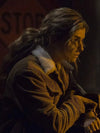 A woman in profile wearing a brown corduroy jacket with a white shearling-style collar, photographed in dim warm lighting against a dark background.