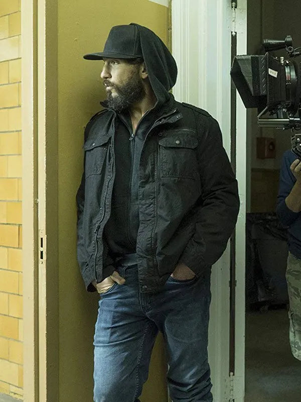 Man in black jacket and cap standing in a doorway with filming equipment in the background