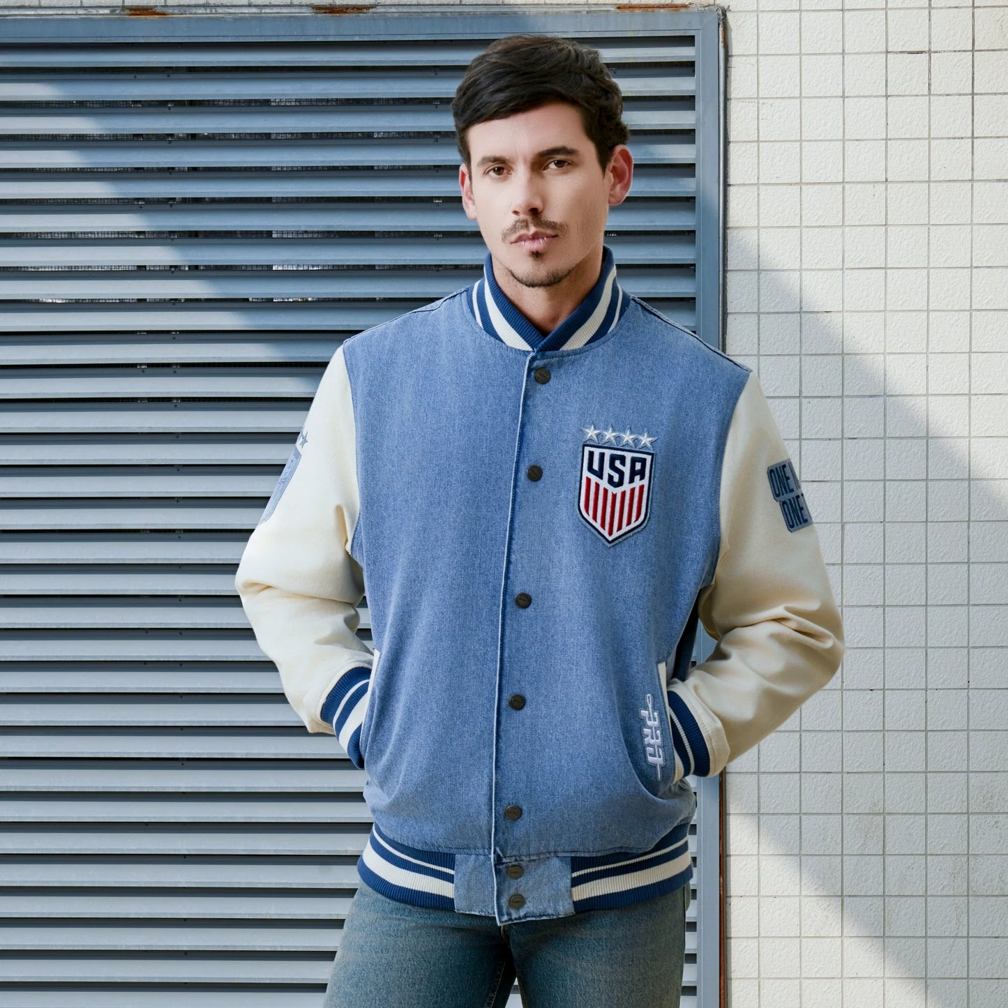 Man wearing a blue and white letterman jacket with USA emblem against a tiled wall.