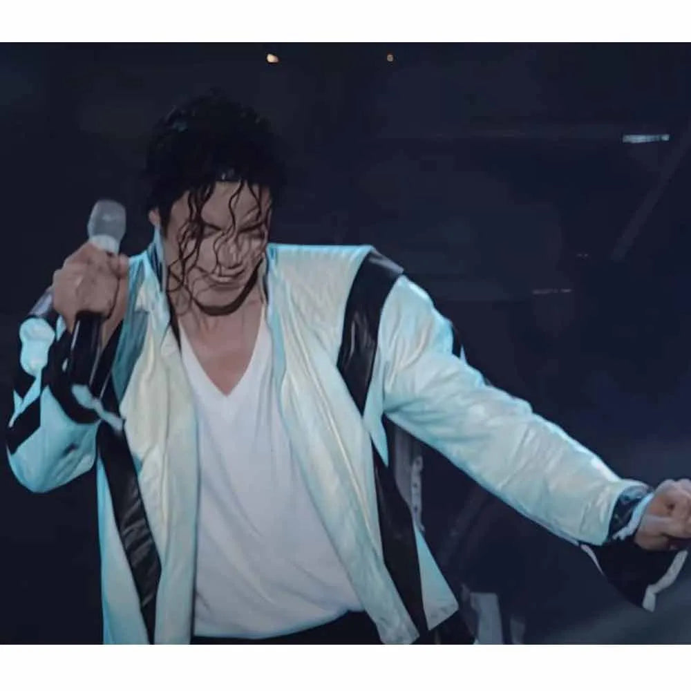 Person performing on stage with a microphone, wearing a light jacket and white shirt.