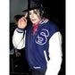 Person wearing a blue and white jacket with a 'S' logo, posing with a finger on their lips.
