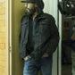 Person wearing a dark jacket and jeans standing in front of a building with a door.