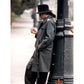 Person wearing a gray trench coat and black hat leaning against a black metal pole on a street.