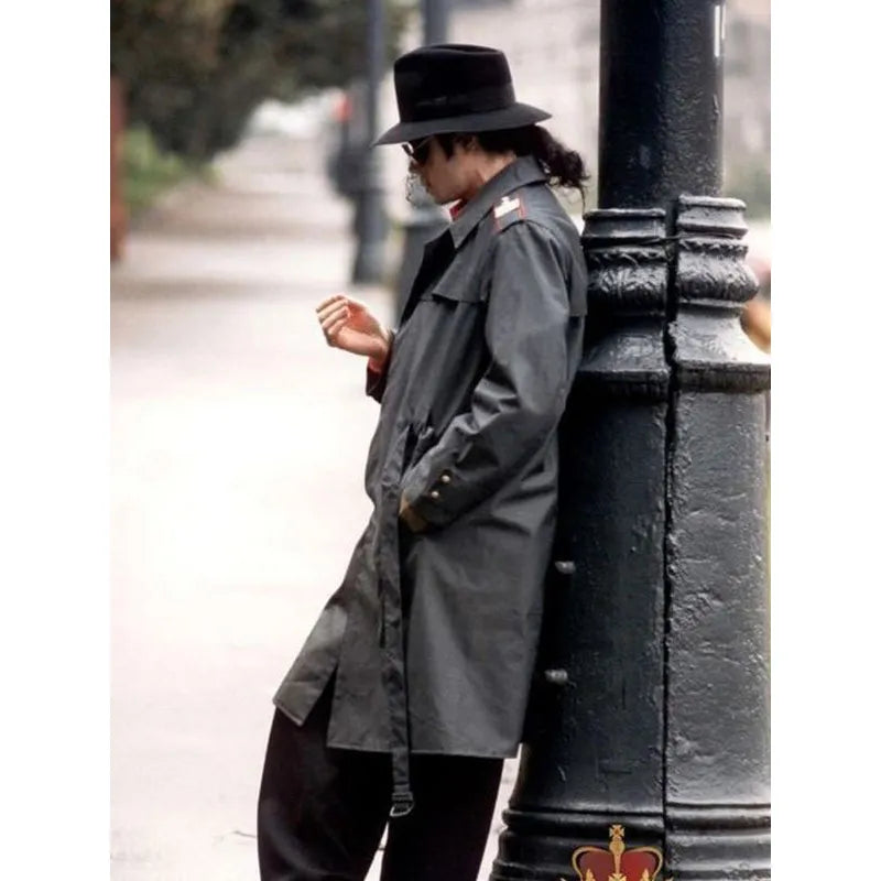 Person wearing a gray trench coat and black hat leaning against a black metal pole on a street.