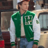 Person wearing a green and red letterman jacket with a logo, standing next to a vintage car.